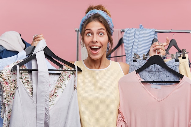 Persone, felicità, shopping, concetto di acquisto. bella donna che ha buon umore mentre tiene in mano molti appendini con i vestiti, provando gioia mentre attende con impazienza un nuovo acquisto o un vestito alla moda