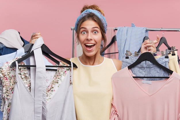 People, happiness, shopping, purchase concept. Beautiful woman having good mood while holding many hangers with clothes, feeling joy while looking forward to new purchase or fashionable outfit