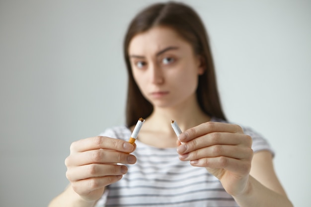 Persone, concetto di cattive abitudini di stile di vita sano. colpo isolato di grave giovane donna bruna posa in studio con sigaretta rotta per pubblicità sociale. ragazza arrabbiata che tiene due metà della sigaretta