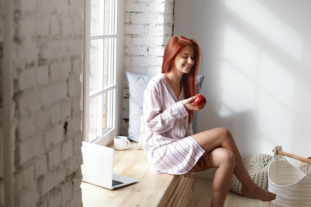 People, healthy lifestyle, food, diet and morning routine concept. Beautiful red haired young Caucasian woman in striped night suit laughing, enjoying calm peaceful morning at home, eating fresh apple