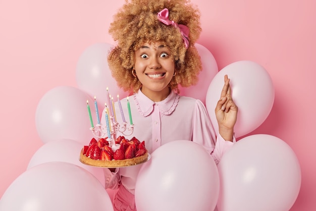 Foto gratuita concetto di festa e celebrazione della gente la donna carina dai capelli ricci vestita con abiti festivi tiene una deliziosa torta di fragole con candele accese tiene le dita incrociate in pose intorno a palloncini gonfiati