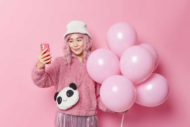 Free photo people and holidays concept. charming asian woman takes selfie via smartphone poses with inflated balloons has fun on party makes memorable photo wears outerwear isolated over pink background