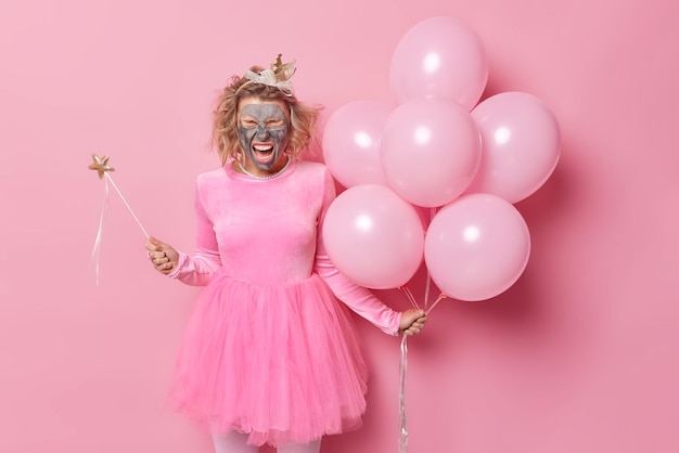 People and holidays concept Positive optimistic young woman with claymask on face excaims loudly hass fun on bachelorette party wears festive clothes holds bunch of inflated helium balloons