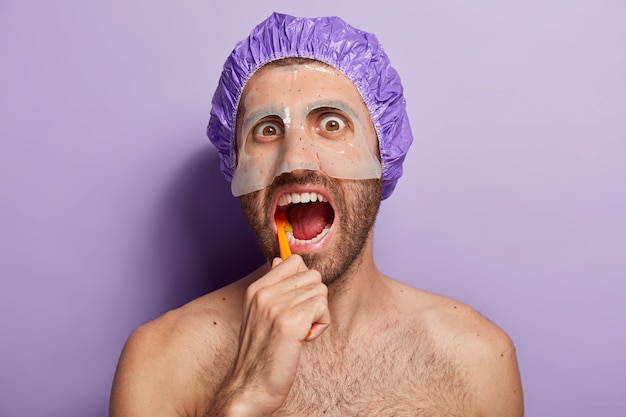 Бесплатное фото Люди, гигиена и концепция утреннего распорядка. крупным планом молодой человек чистит зубы зубной щеткой, держит рот открытым, носит банный колпак, маску красоты на лице