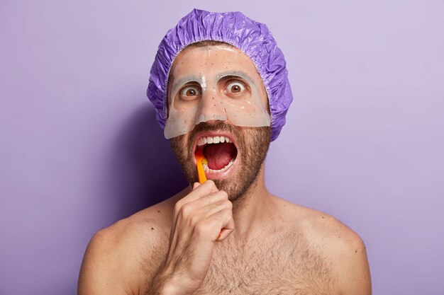 Люди, гигиена и концепция утреннего распорядка. Крупным планом молодой человек чистит зубы зубной щеткой, держит рот открытым, носит банный колпак, маску красоты на лице