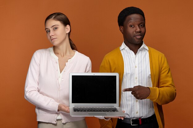 People, job, modern technology and communication concept. Successful attractive young European woman and her stylish African male colleague posing together in , holding laptop with black display