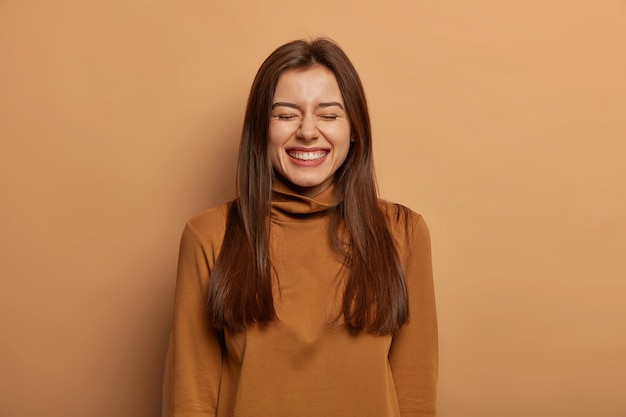 People and joy concept. Overjoyed dark haired adult woman laughs happily with closed eyes, talks casually with friend, cannot hold laughter, wears casual turtneck, isolated on brown wall