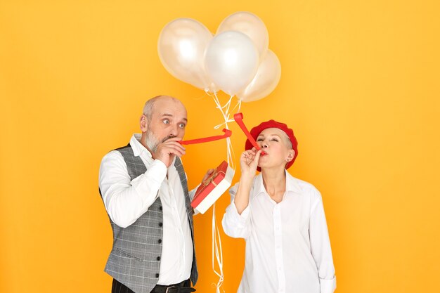 People, joy, fun and celebration concept. Portrait of elegant bald male pensioner and beautiful mature woman enjoying birthday party, blowing whistle