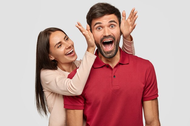 People, joy, pleasant moment in life. Overjoyed brunette European young female stands near her boyfriend, going to cover eyes and make surprise, have fun together, isolated over white wall.