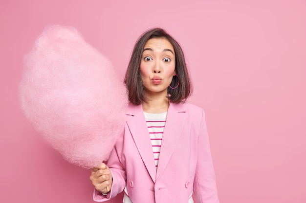 People and leisure concept. Lovely brunette Asian woman with dark hair keeps lips folded holds yummy sweet candy floss on stick dressed in stylish clothes isolated over pink  wall.