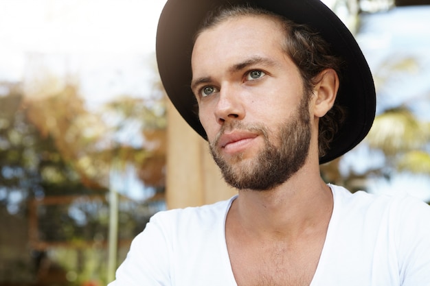 People and leisure. Headshot of fashionable young male with trendy beard wearing black hat having thoughtful expression, looking into distance , planning day