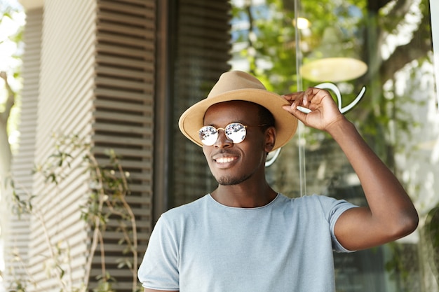 People, leisure and lifestyle concept. Happy and relaxed young black European man in stylish clothing adjusting brims of his hat smiling broadly while flirting with pretty woman at sidewalk cafe