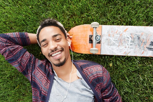 People, leisure and lifestyle concept. Smiling joyful skater rests after training, lies on green grass near skateboard