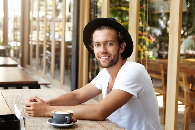 People, leisure and lifestyle concept. Successful young man in black hat and casual t-shirt having coffee sitting at sidewalk restaurant