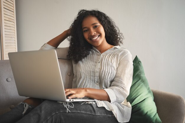 People, leisure, modern lifestyle, technology and electronic gadgets concept. Attractive happy young mixed race woman enjoying online communication, having video chat using laptop computer at home