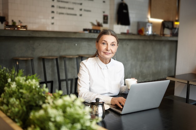 Free photo people, leisure and modern technologies concept. picture of blue eyed elderly lady sitting at cafe table in front of open laptop computer, using wireless internet connection and drinking coffee