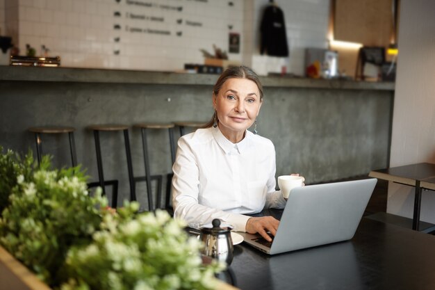 People, leisure and modern technologies concept. Picture of blue eyed elderly lady sitting at cafe table in front of open laptop computer, using wireless internet connection and drinking coffee