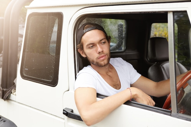 People, leisure, travel and vacations concept. Fashionable unshaven man wearing snapback driving his white crossover utility vehicle while trying to get around traffic jam