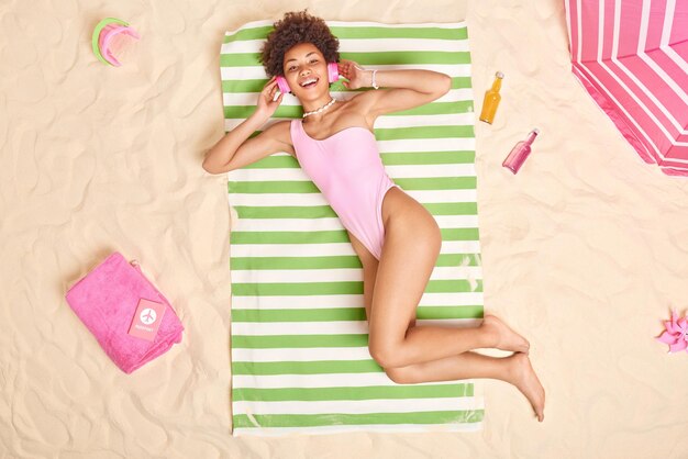 People leisure and vacation concept Cheerful curly haired beautiful woman dressed in swimwear enjoys favorite playlist while lying in sun at beach has lazy day unforgettable summer holidays