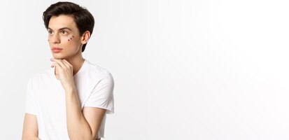 People lgbtq and beauty concept beautiful androgynous man with glitter under eyes looking at upper left corner posing against white background