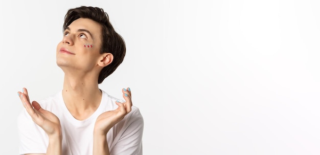 People lgbtq and beauty concept Closeup of handsome androgynous man with glitter on face and polished fingernails looking dreamy up and smiling white background