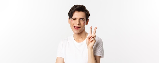 People lgbtq community and lifestyle concept happy and cute gay man with glitter on face showing peace sign and smiling celebrating pride holiday white background