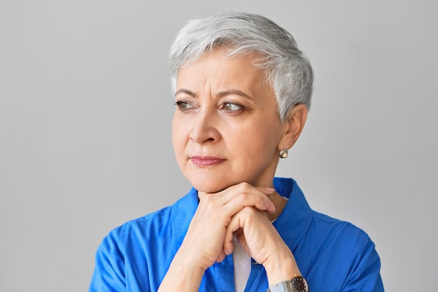 People, lifestyle and age concept. Serious Caucasian woman pensioner with gray pixie hair placing chin on clasped hands and looking away, feeling upset and lonely. Portrait of sad mature female