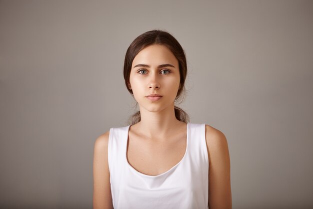 People, lifestyle, beauty and fashion concept. Close up portrait of beautiful stylish young European female posing isolated having calm peaceful facial expression wearing white sleeveless top