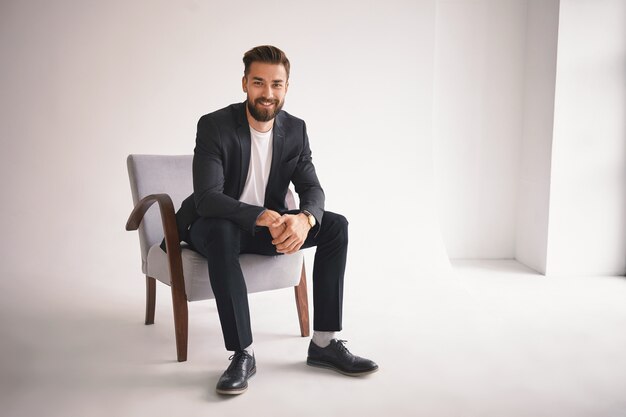 People, lifestyle, business, style, fashion and men's wear concept. Positive successful young CEO sitting in armchair, smiling, dressed in elegant shoes, trousers, jacket and white t-shirt