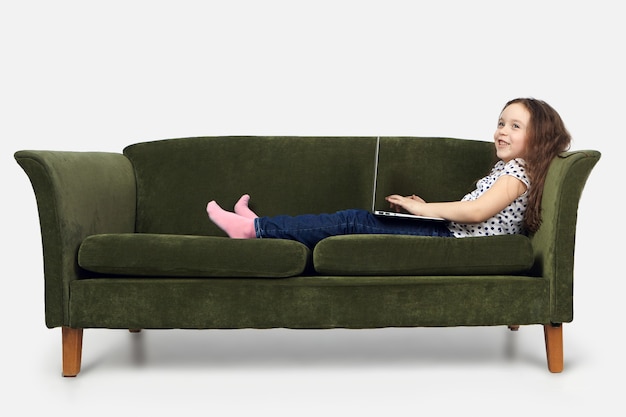 People, lifestyle, childhood and modern technology concept. Pretty schoolgirl relaxing at home with laptop after classes at school, watching cartoons or video blog, looking away with happy smile