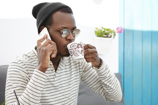 People, lifestyle, communication and modern technology concept. Attractive young Afro American student having phone conversation while drinking tea or coffee