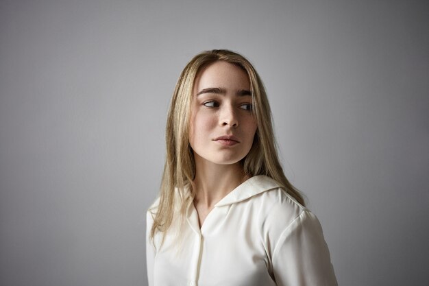 People and lifestyle concept. Close up image of blonde freckled girl in her twenties posing in studio. Attractive stylish young female wearing white silk blouse, looking away with curious expression