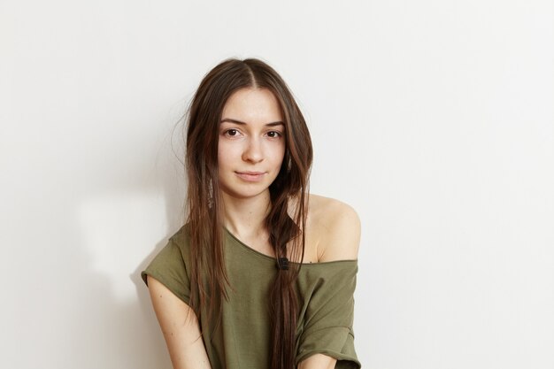 People and lifestyle concept. Headshot of beautiful Caucasian brunette teenage woman with wild hairstyle wearing trendy army green t-shirt with one shoulder showing up, posing isolated at white wall
