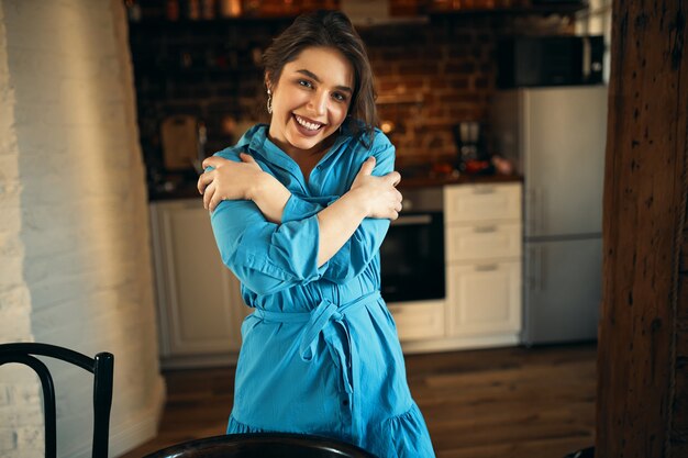 人とライフスタイルのコンセプト。彼女の胸に腕を組んで、幸せそうな表情を持って、彼女の胸に腕を組んでキッチンのインテリアの背景に立っている魅力的な笑顔で魅力的なかわいい若い女性の屋内ショット