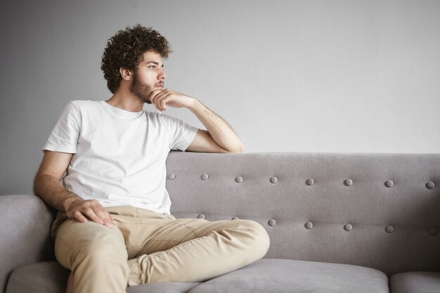 People and lifestyle concept. Picture of handsome serious young bearded guy having pensive facial expression, touching his stubble, deep in thoughts, pondering, searching for solution to problem