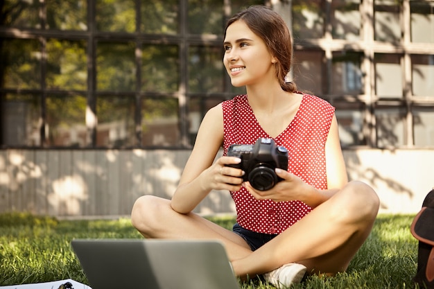 Persone e concetto di stile di vita. immagine del fotografo professionista femminile in vestiti alla moda che tiene la fotocamera dslr seduto sull'erba davanti al computer portatile, ritoccando le foto del servizio fotografico estivo