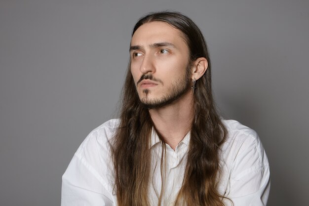 People and lifestyle concept. Picture of trendy looking young creative man with long hair and beard posing indoors in white shirt, having pensive concentrated look, working or making decision