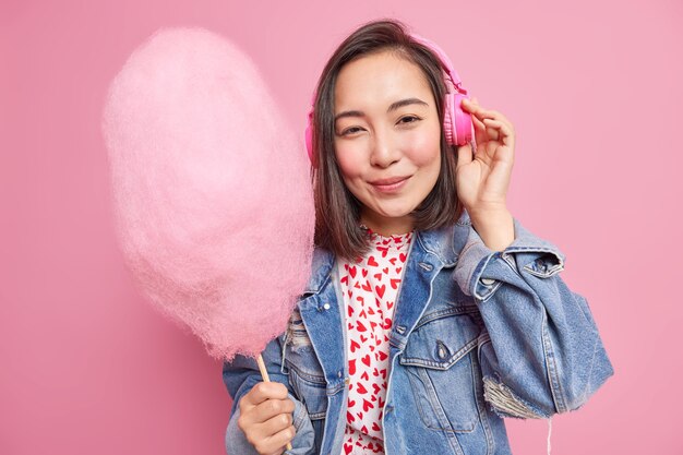 사람과 라이프 스타일 개념. 예쁜 아시아 십대 소녀는 헤드폰을 통해 음악을 듣고 유행 데님 재킷을 입은 맛있는 솜사탕을 보유하고 분홍색 벽에 고립 된 자유 시간을 즐깁니다.