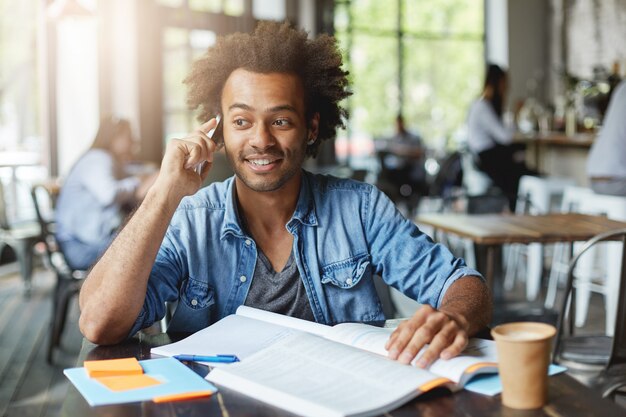 People, lifestyle, education and modern technology concept. Candid shot of cheerful Afro American male student in stylish wear enjoying nice conversation on cell phone while doing homework at canteen