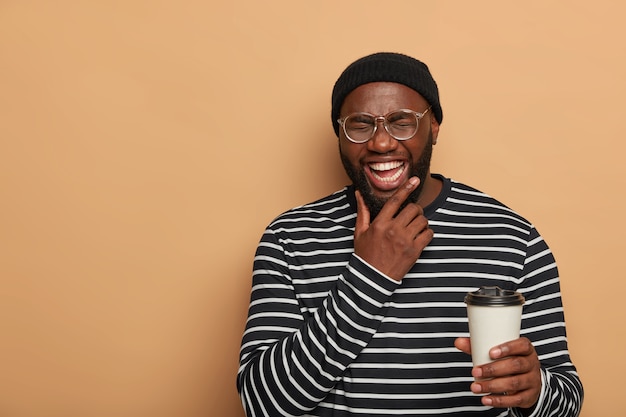 People, lifestyle, emotions concept. joyful black man holds chin, smiles sincerely, drinks takeout coffee