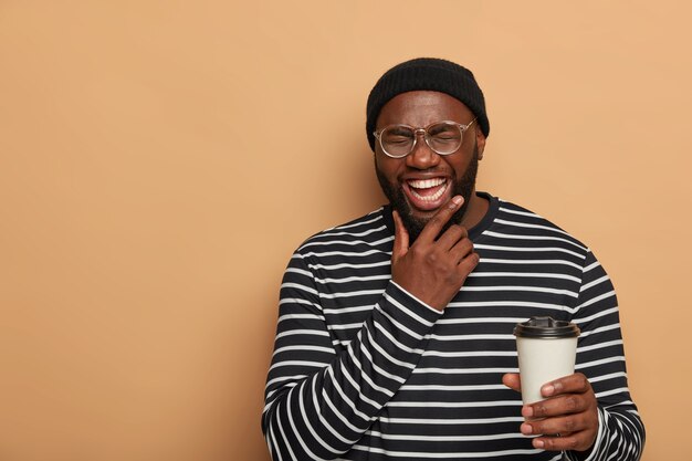 People, lifestyle, emotions concept. Joyful black man holds chin, smiles sincerely, drinks takeout coffee