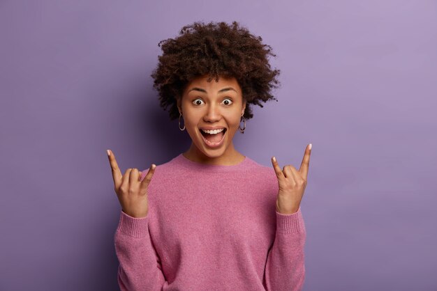 人、ライフスタイル、感情、感情の概念。興奮した感情的な女性のロックファンは素晴らしい夏のキャンパスパーティーを楽しんで、楽しんで、ヘビーメタルのサインを見せ、のんきな屋内に立って、積極的にジェスチャーをします