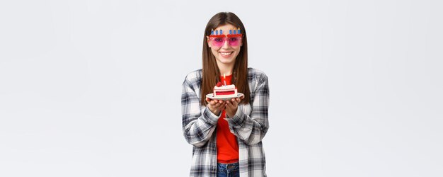 Люди, образ жизни, праздники и празднование эмоций, концепция Веселая милая девушка в очках, держащая торт ко дню рождения, празднует день рождения, загадывая желание задуть зажженную свечу на белом фоне