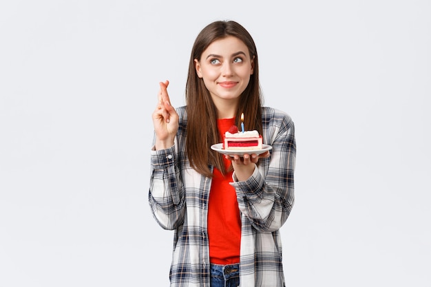 People lifestyle, holidays and celebration, emotions concept. Dreamy hopeful birthday girl in casual clothes, cross fingers and look up making wish, holding b-day cake, blowing candle.