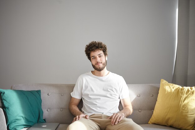 People, lifestyle, interior and design concept. Cheerful attractive young Caucasian man with stubble and stylish wavy hairdo sitting on comfortable couch with decorative pillows and smiling