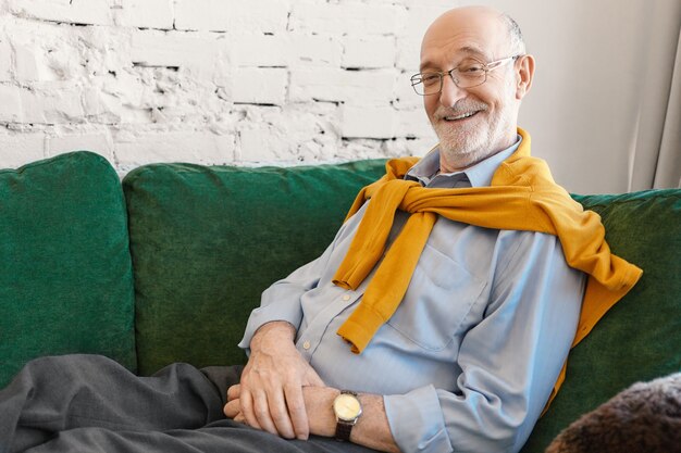 People, lifestyle, joy, rest and relaxation concept. Horizontal shot of handsome emotional 70 year old grandfather wearing elegant clothes and spectacles relaxing at home on couch, smiling broadly