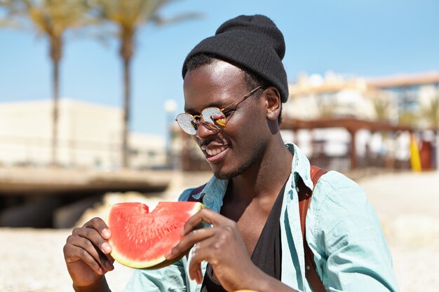 Люди, образ жизни, досуг, каникулы и концепция путешествия. Счастливый расслабленный афроамериканский мужчина-турист держит в руках кусочек сочного арбуза, наслаждаясь сладкими и спелыми фруктами в солнечный летний день