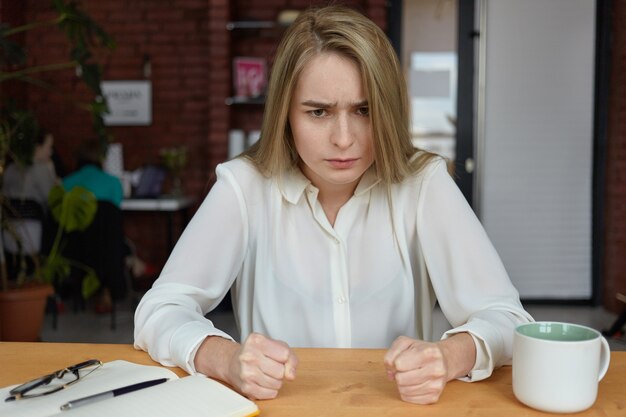 People, lifestyle and negative human emotions. Furious young businesswoman in white blouse working while sitting at cafeteria during coffee break, being angry because her lunch is not ready yet