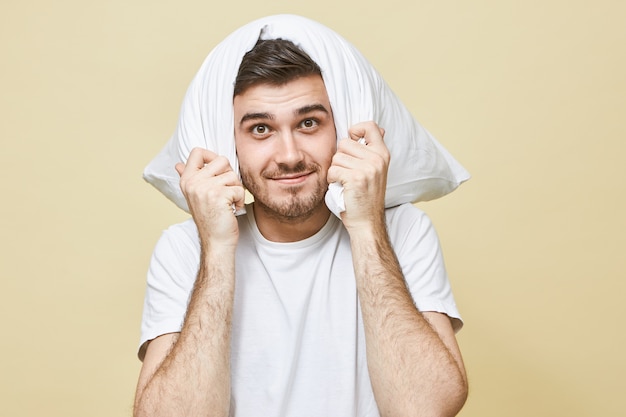 People, lifestyle, rest, health and recreation concept. Handsome unshaven young guy in white pajamas covering ears with pillow, smiling, because not hearing snore coming from his asleep wife