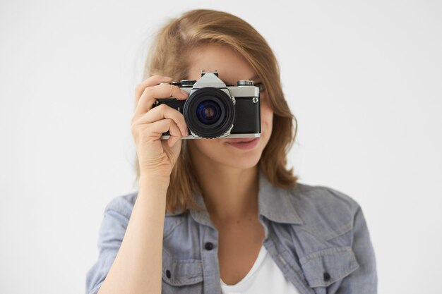 人、ライフスタイル、テクノロジーのコンセプト。ロールフィルムカメラを顔に持って、あなたの写真を撮るスタイリッシュな女の子のスタジオショット。写真を撮るためにビンテージデバイスを使用して若い女性の写真家